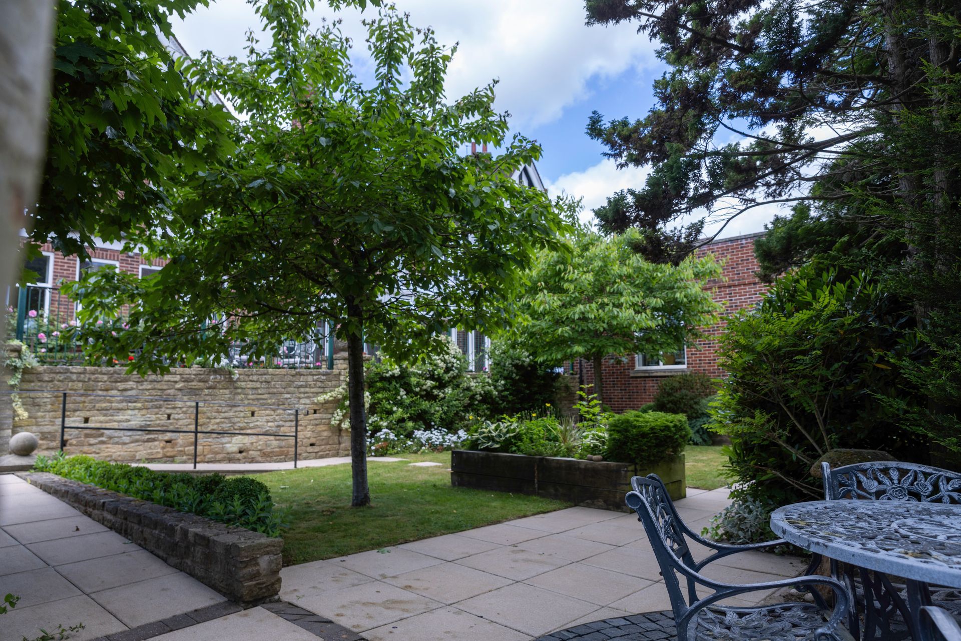A garden with a patio, trees, and a brick building in the background. Sunny day with blue sky.