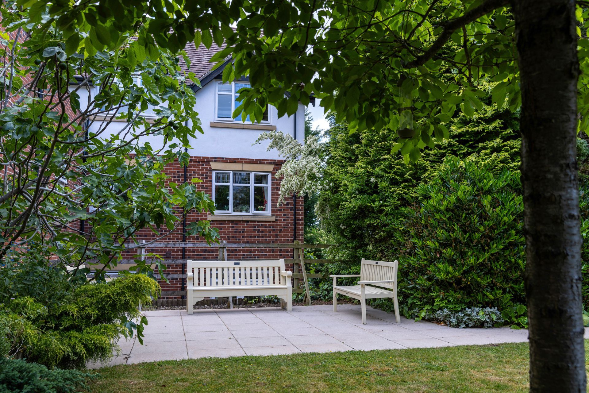 A brick house with white trim and a patio with two benches surrounded by green trees and bushes.