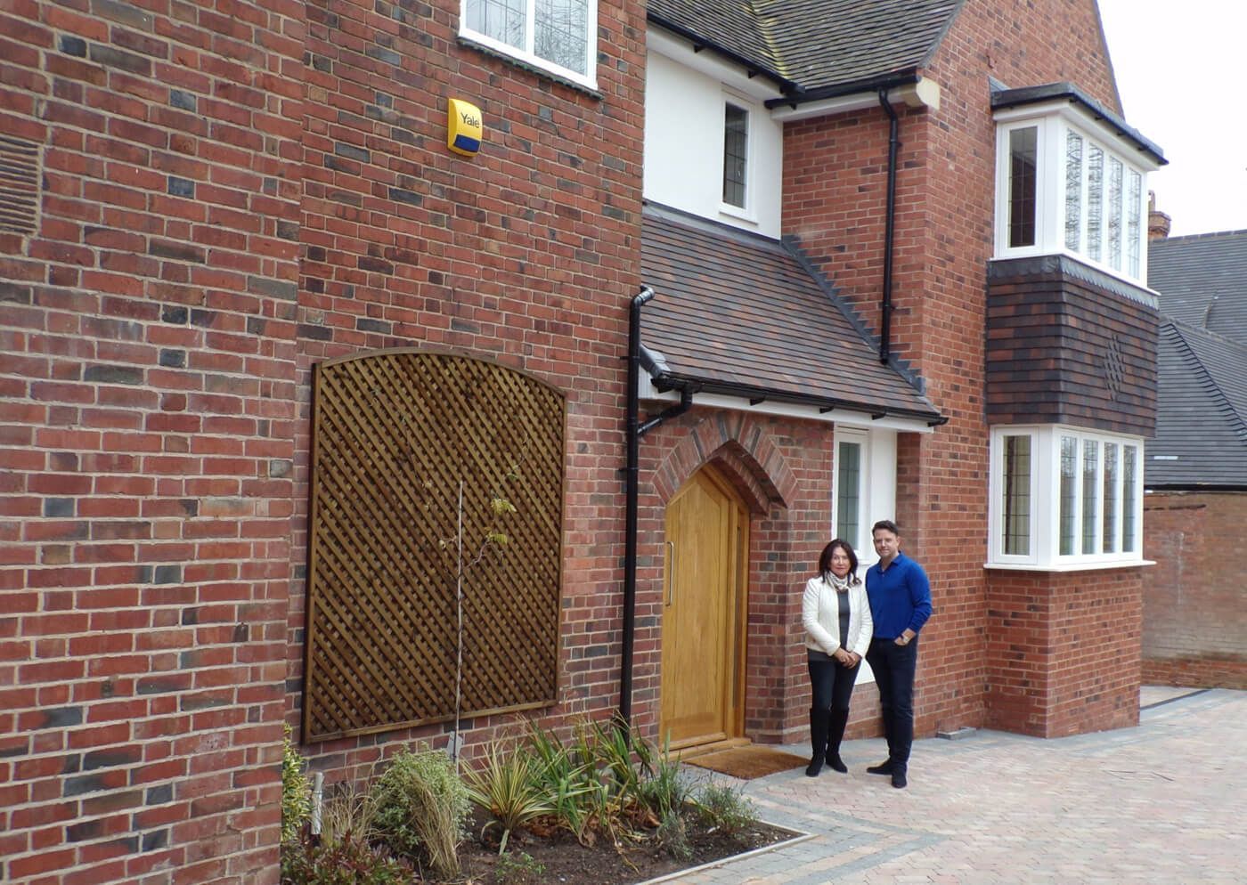 A couple stands in front of a brick home with a wooden door and bay window.