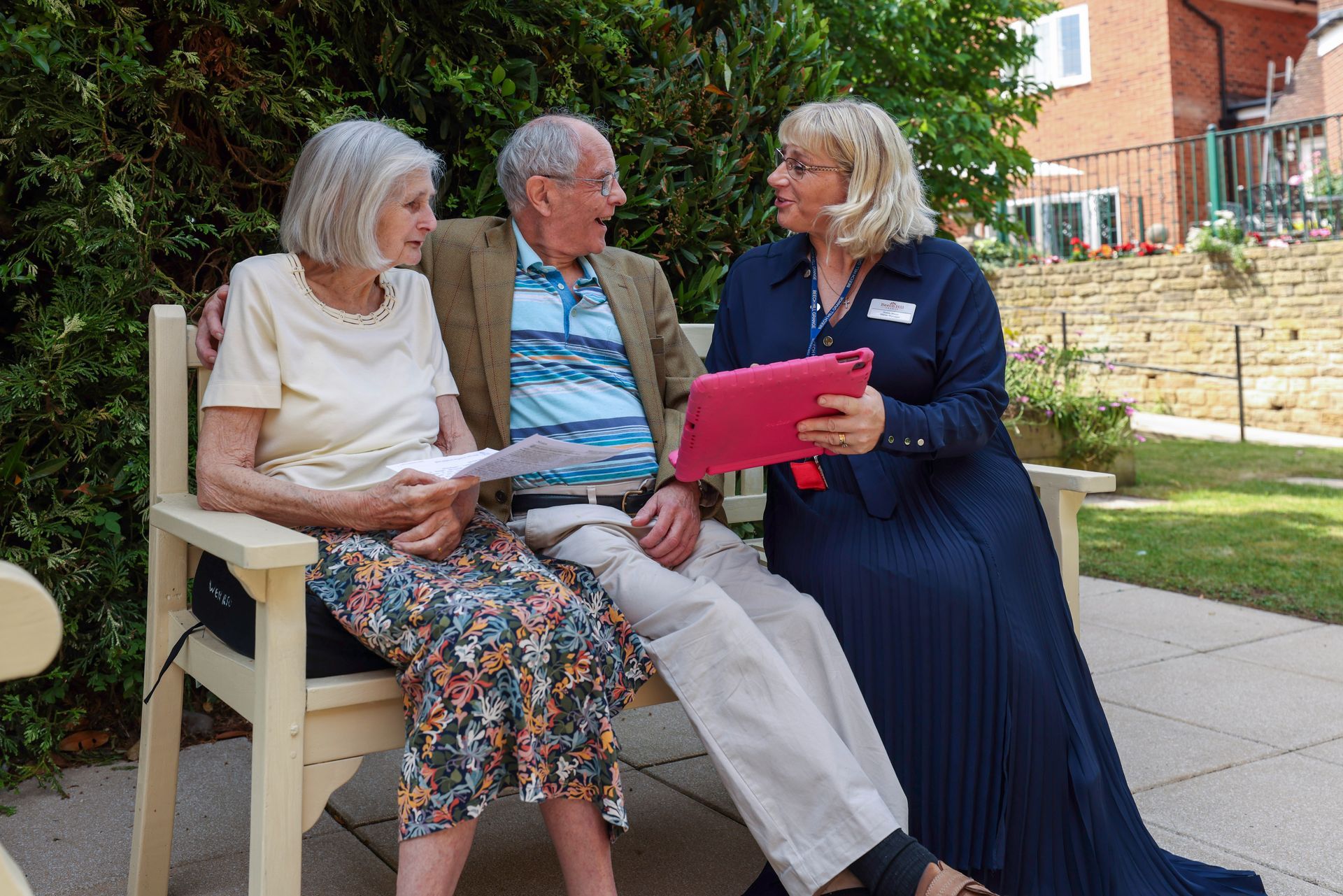 An older couple sits on a bench, speaking with a woman holding a tablet, outdoors.
