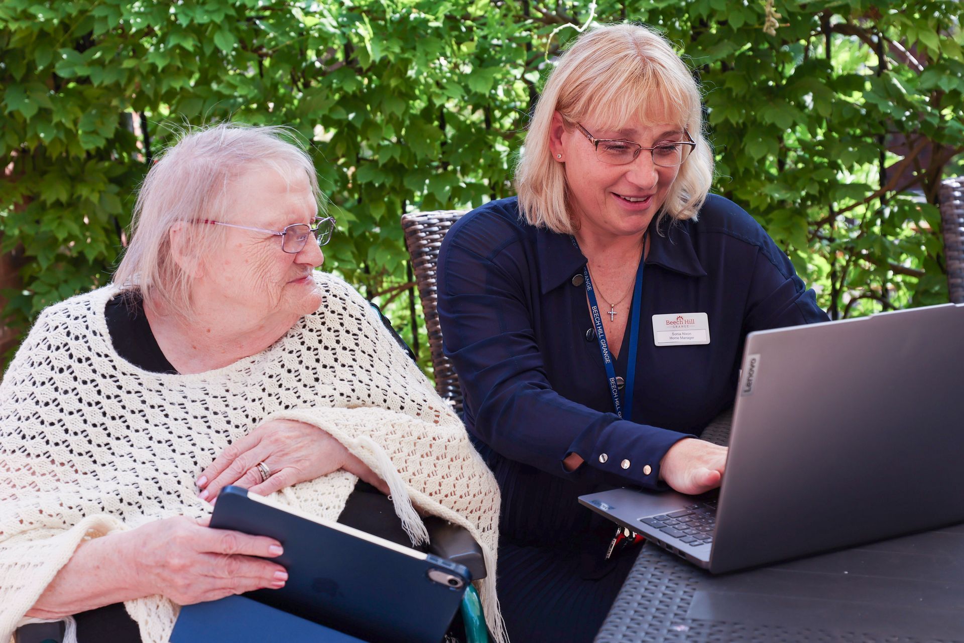 An older woman and a younger woman look at a laptop outdoors; the younger woman points, and the older woman holds a notebook.
