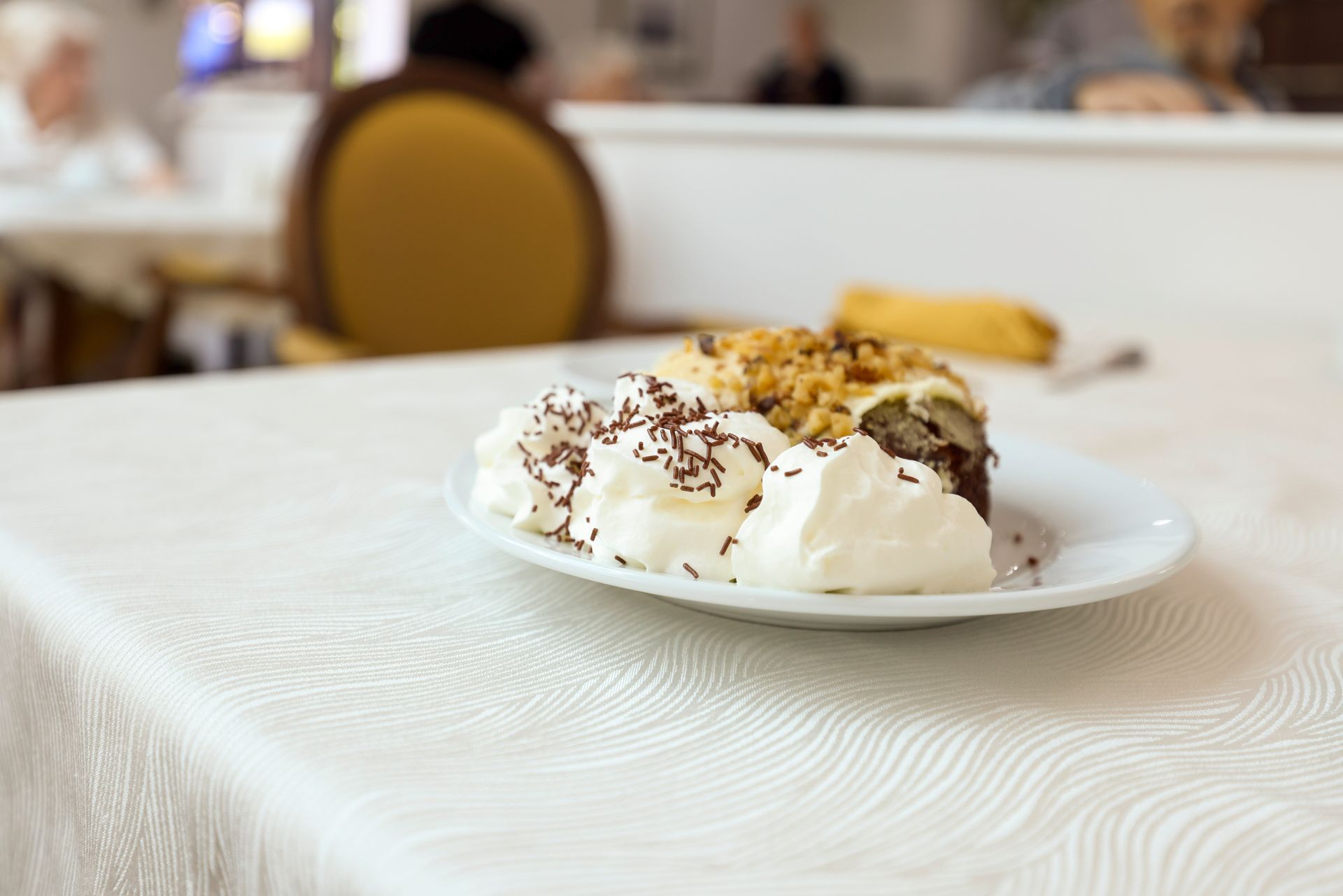 Cake slice with whipped cream and chocolate sprinkles on a white plate, table setting.