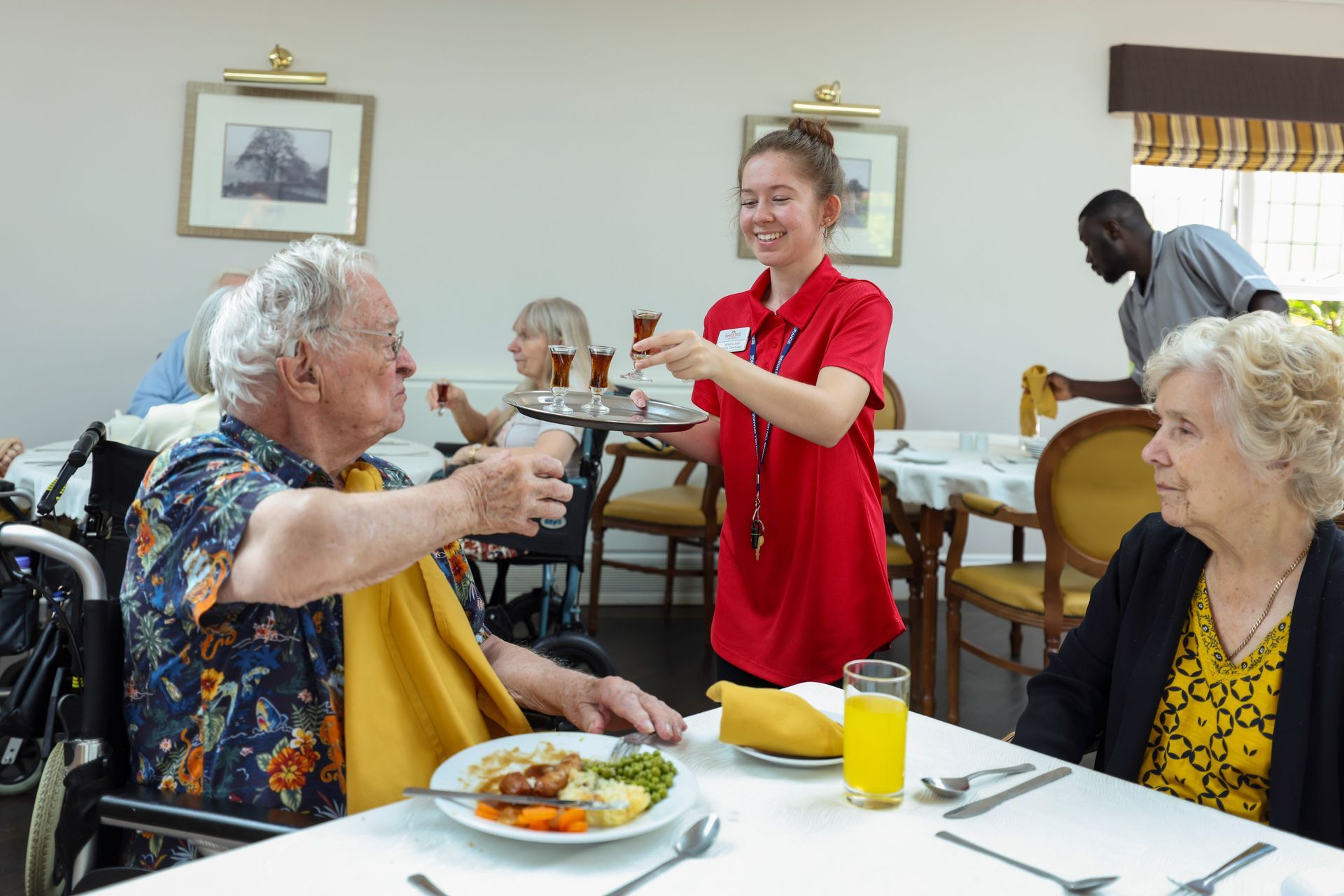 A young woman in red serves drinks on a tray to elderly residents at a dining table. Another man is clearing the table.