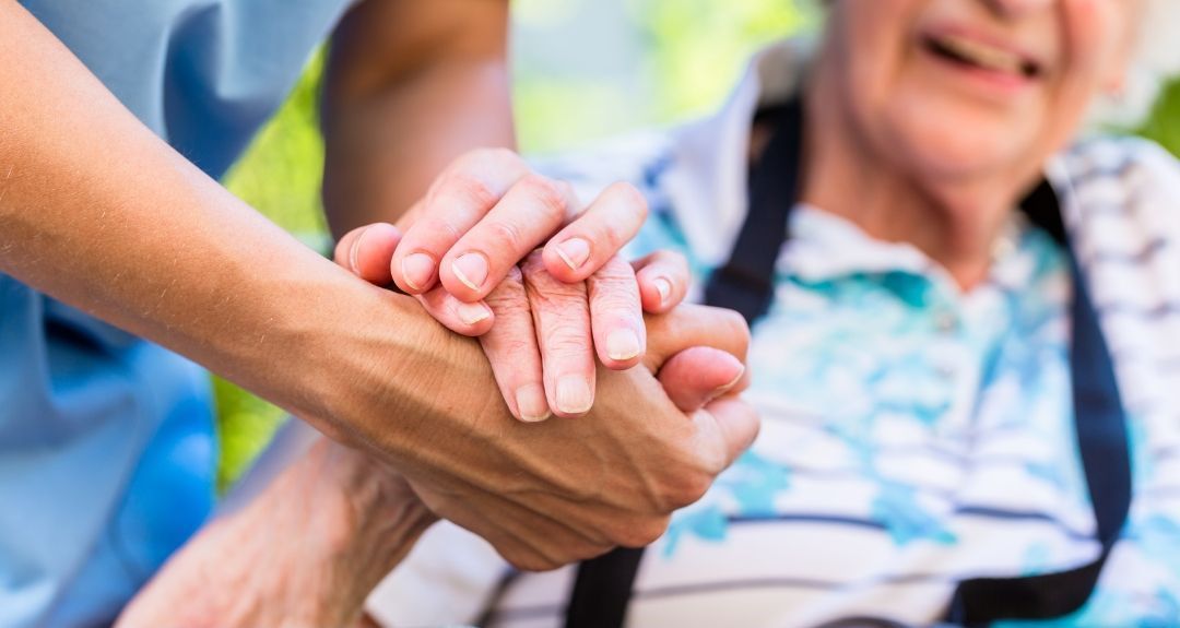 Caregiver holding the hand of an elderly woman. Both are outdoors, the woman is smiling.
