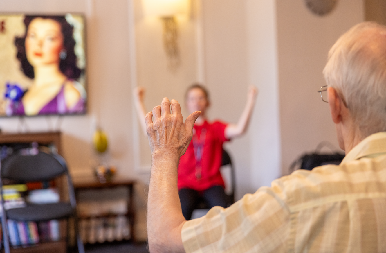An older man stretches his arms, mirroring an instructor in a red shirt, indoor setting, focused.