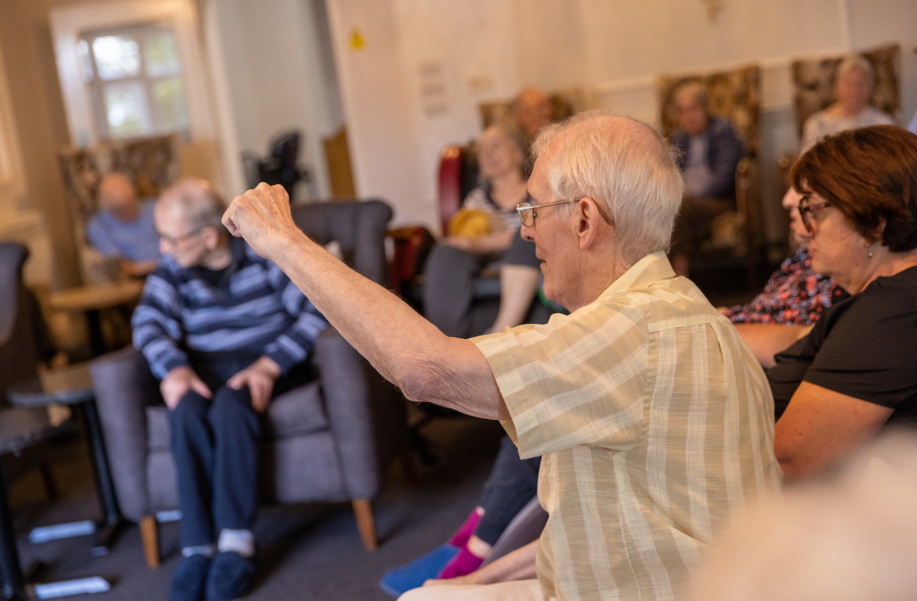 Elderly man raises arm, speaking in a group setting with others seated in chairs indoors.