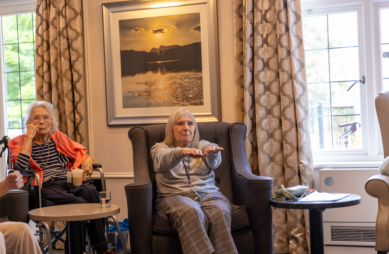 Two women in a living room; one exercises arms, the other watches. A painting and window are in the background.