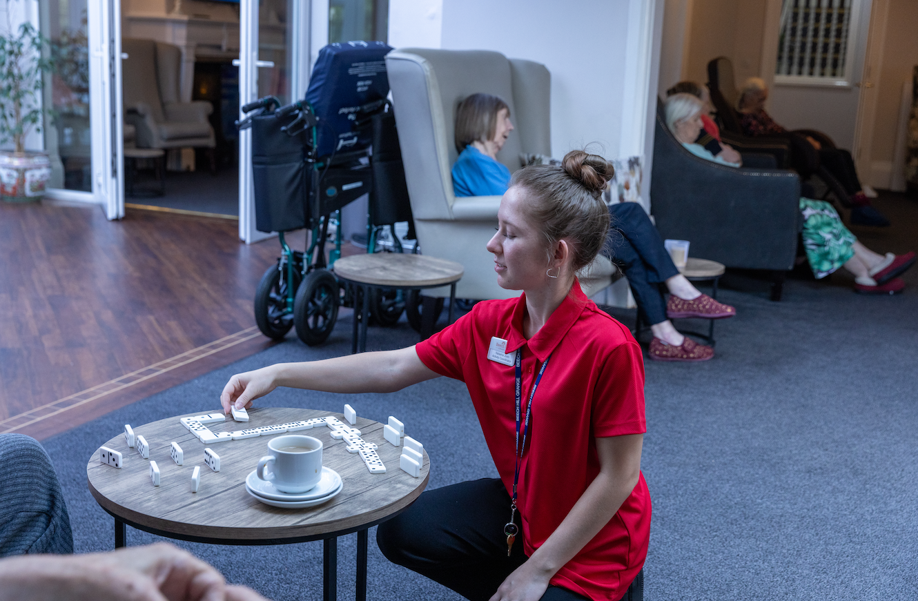 Woman in red shirt plays dominoes on a small table with a cup of coffee. Other people relax in armchairs in the background.