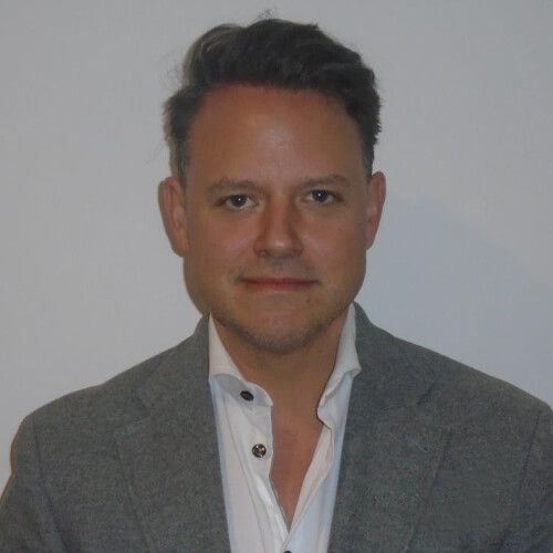 A man in a gray blazer and white shirt looking at the camera against a plain background.