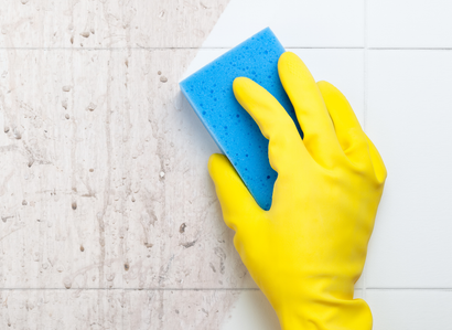 A hand in a yellow rubber glove uses a blue sponge to wipe grime off white wall tiles.