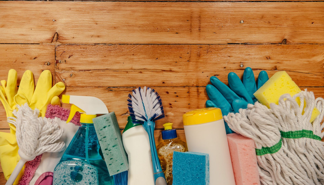 Cleaning supplies including rubber gloves, sponges, a brush, mop heads, and bottles arranged on a wooden surface.