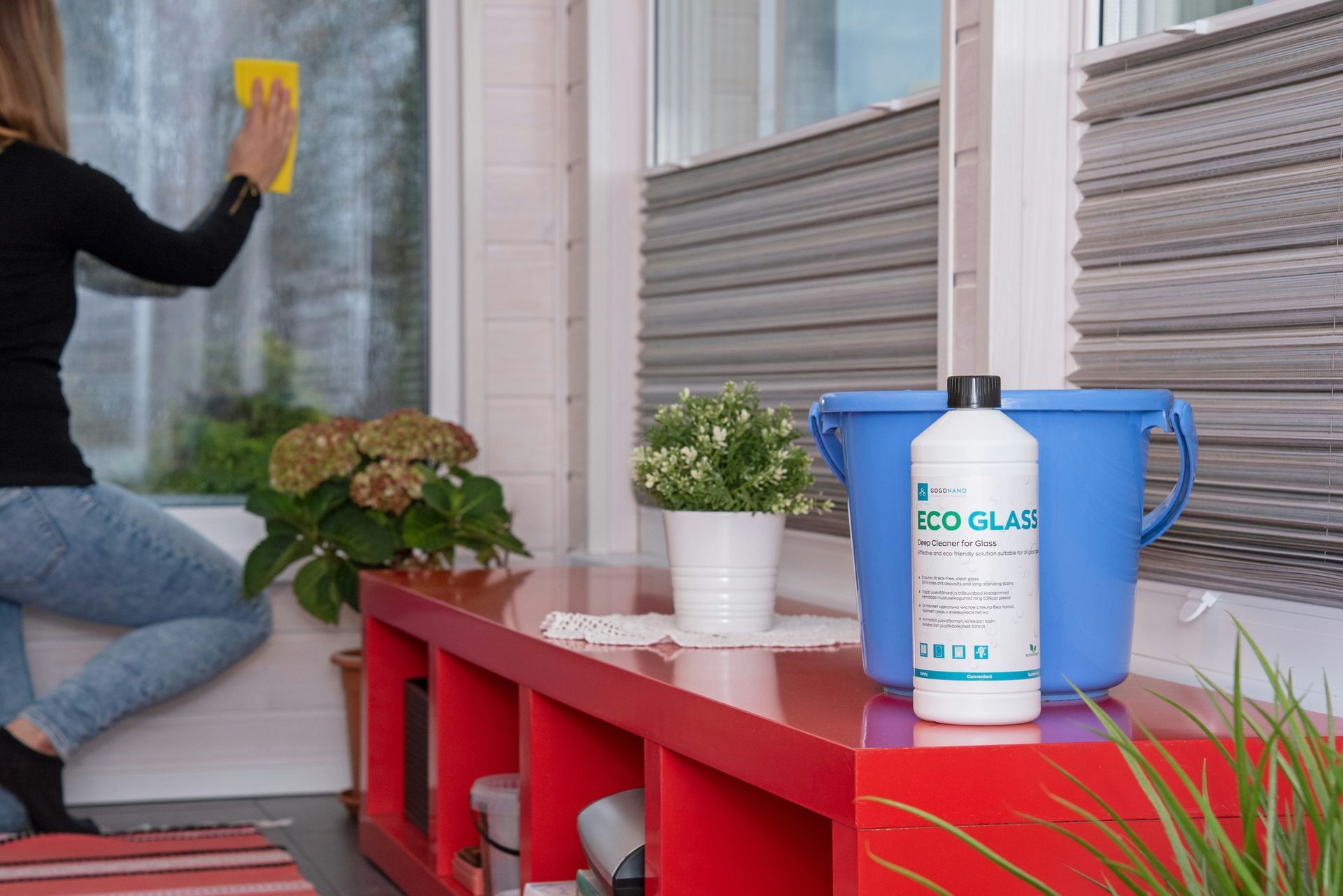 A person wipes a window near a red shelf holding a potted plant, a blue bucket, and a cleaning solution bottle.