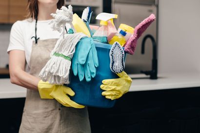 Person in an apron and yellow gloves holding a blue bucket filled with cleaning supplies in a kitchen.