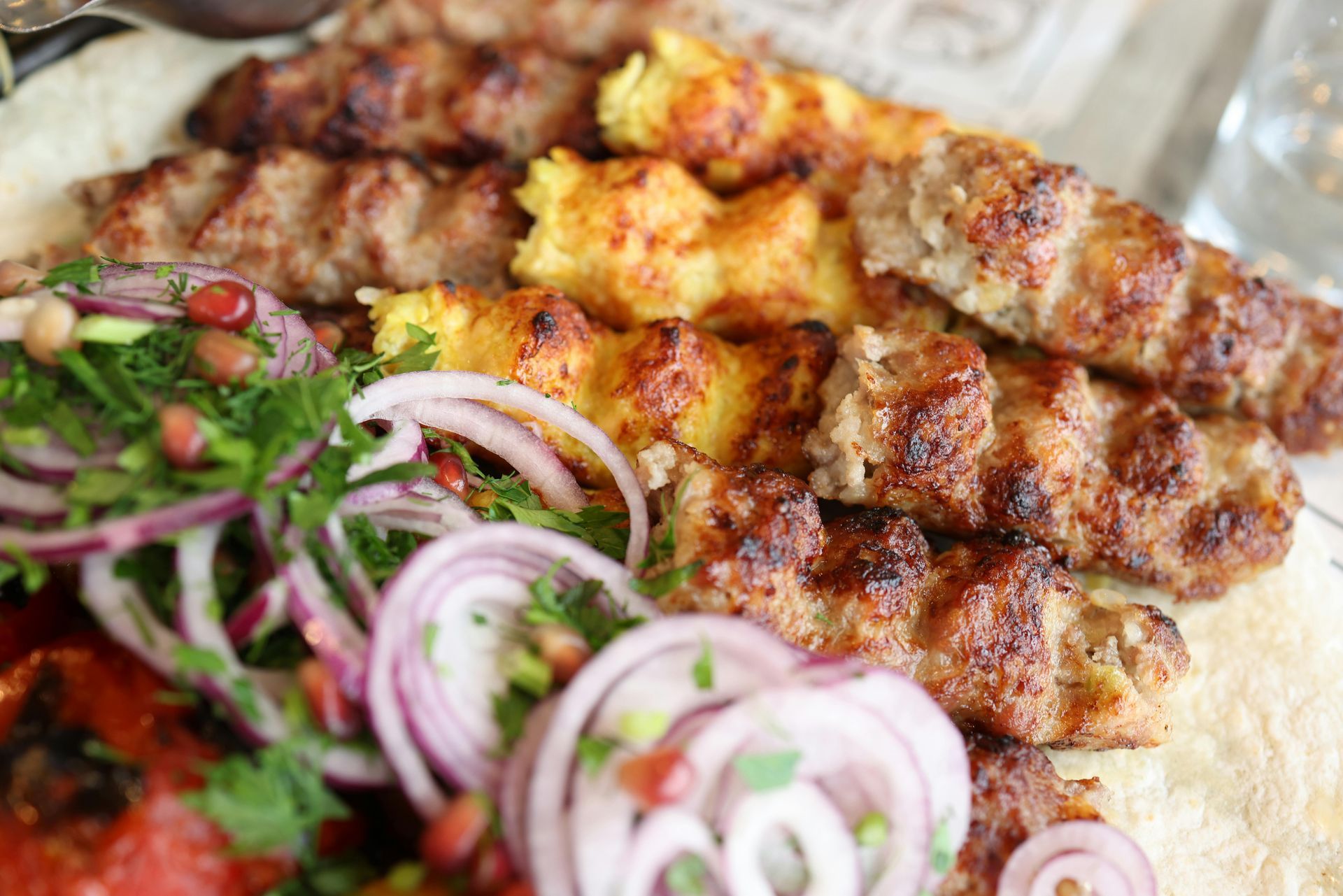 Un primo piano di spiedini grigliati serviti su pane piatto con contorno di cipolle rosse a fette, erbe aromatiche fresche e pomodori grigliati.