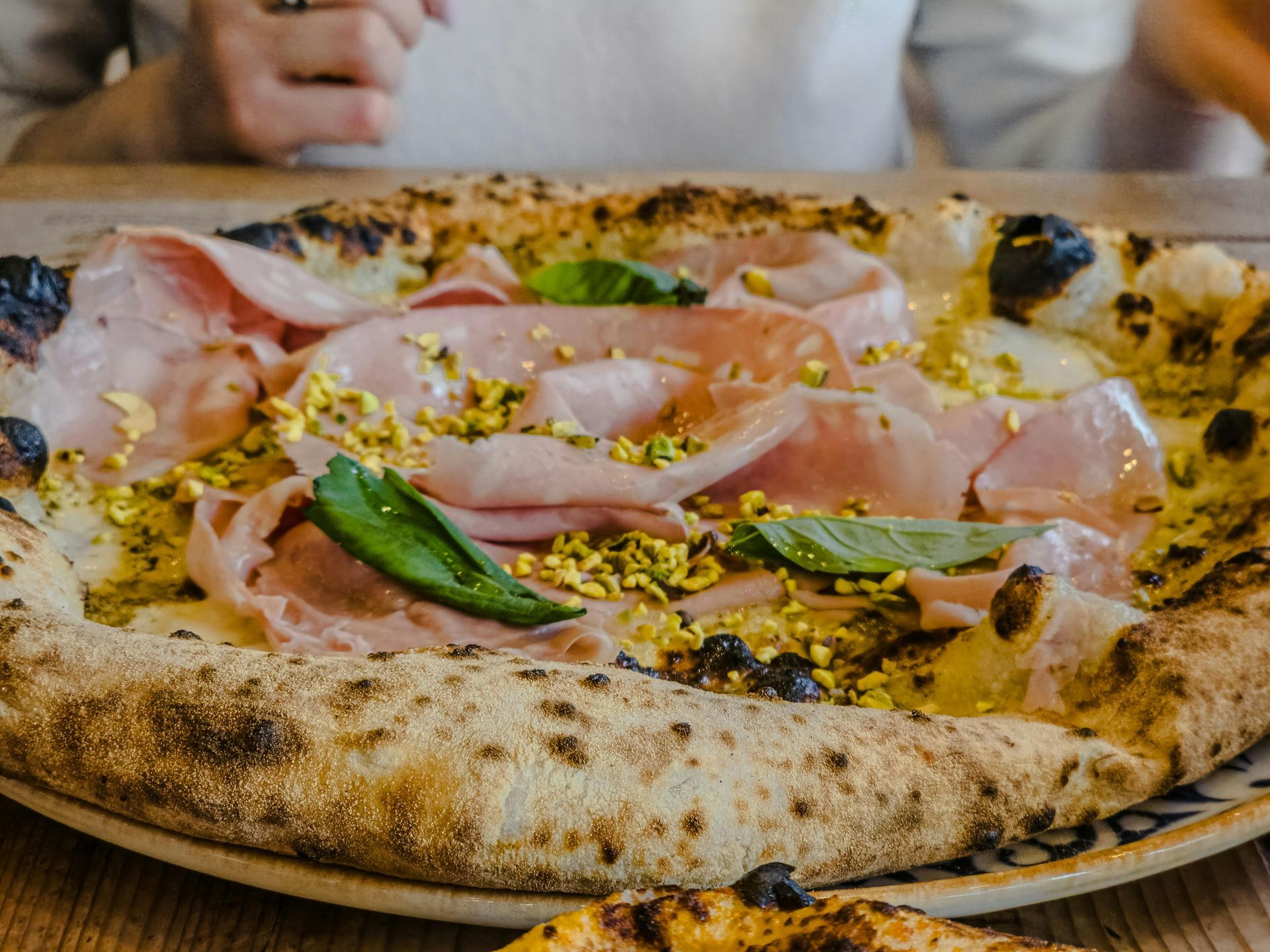 Una pizza gourmet condita con mortadella ripiegata, pistacchi tritati e foglie di basilico fresco, servita su un piatto rustico.