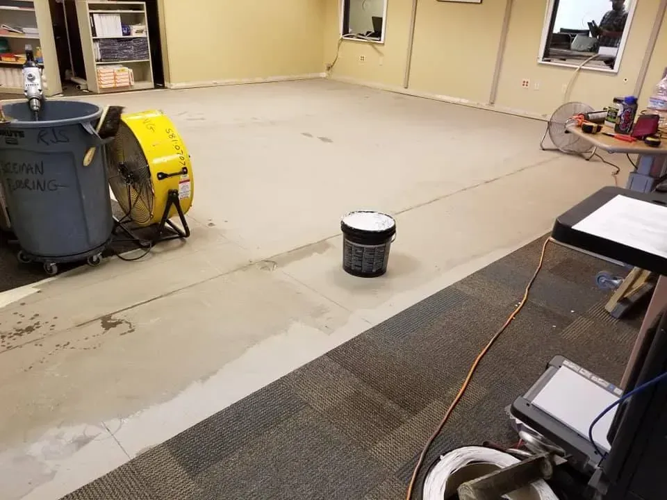 A room with a bucket of paint and a fan on the floor.