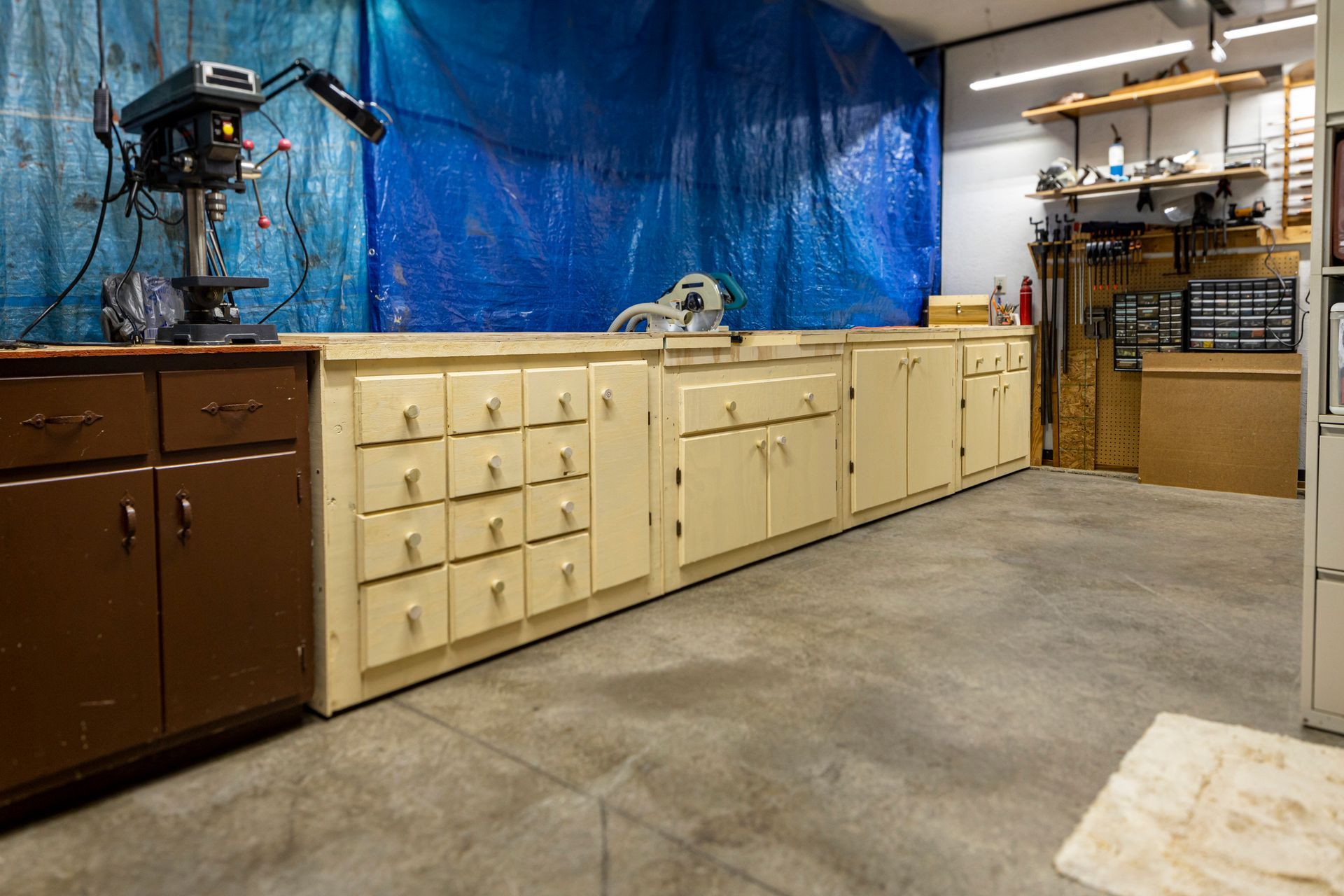 Workshop with tan and brown cabinets, a drill press, and a blue tarp backdrop.