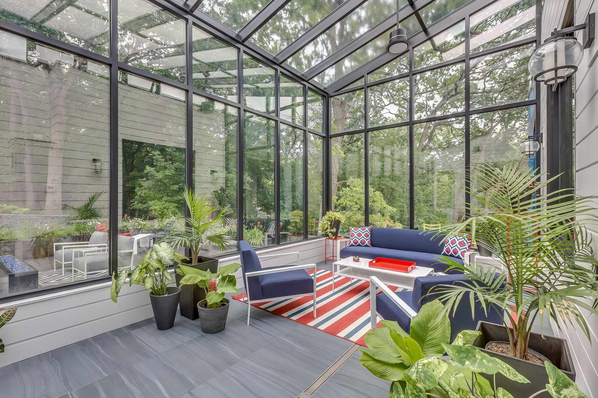 Sunroom with glass walls, blue furniture, red rug, and potted plants, overlooking a wooded area.