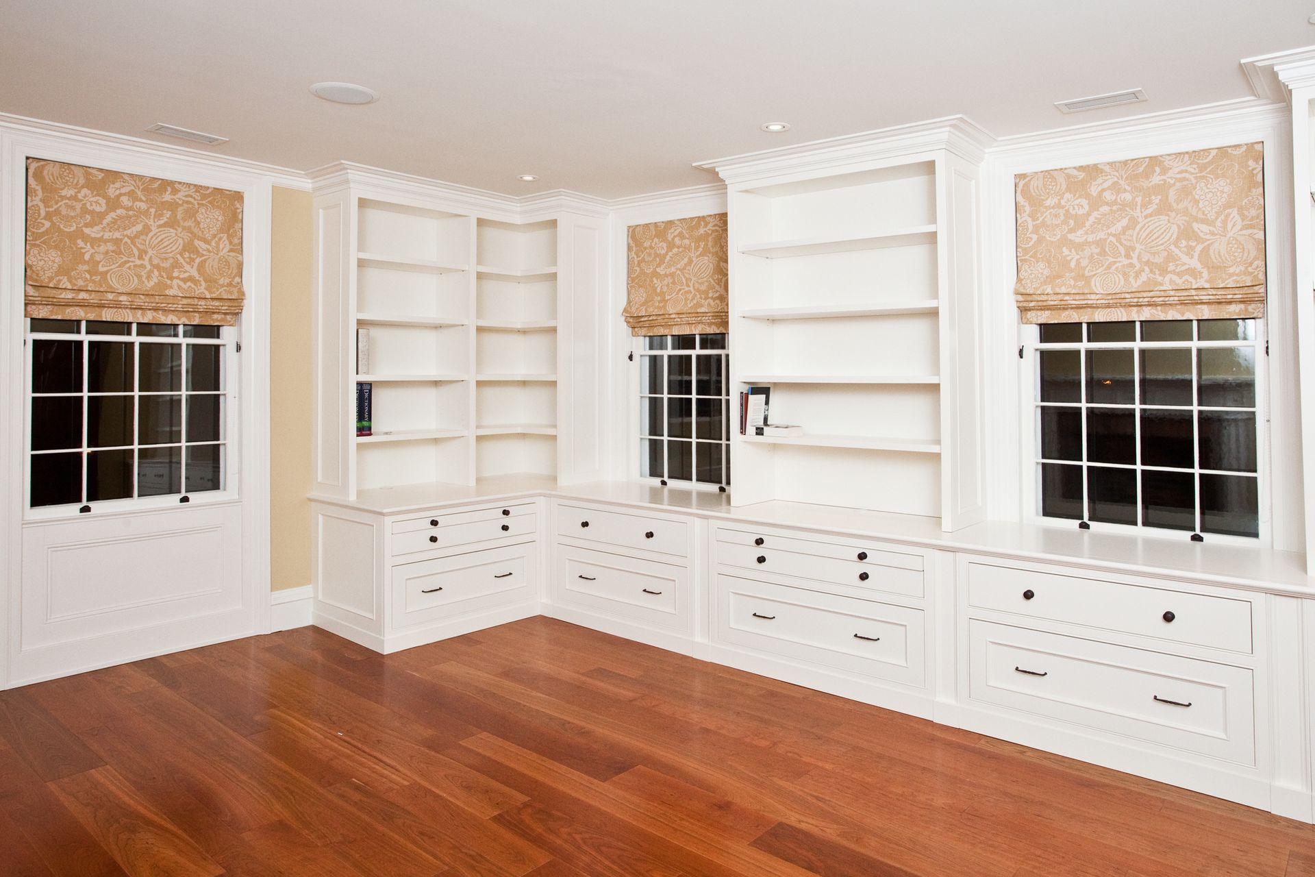 Built-in white shelves and drawers around windows with patterned shades, hardwood floor.