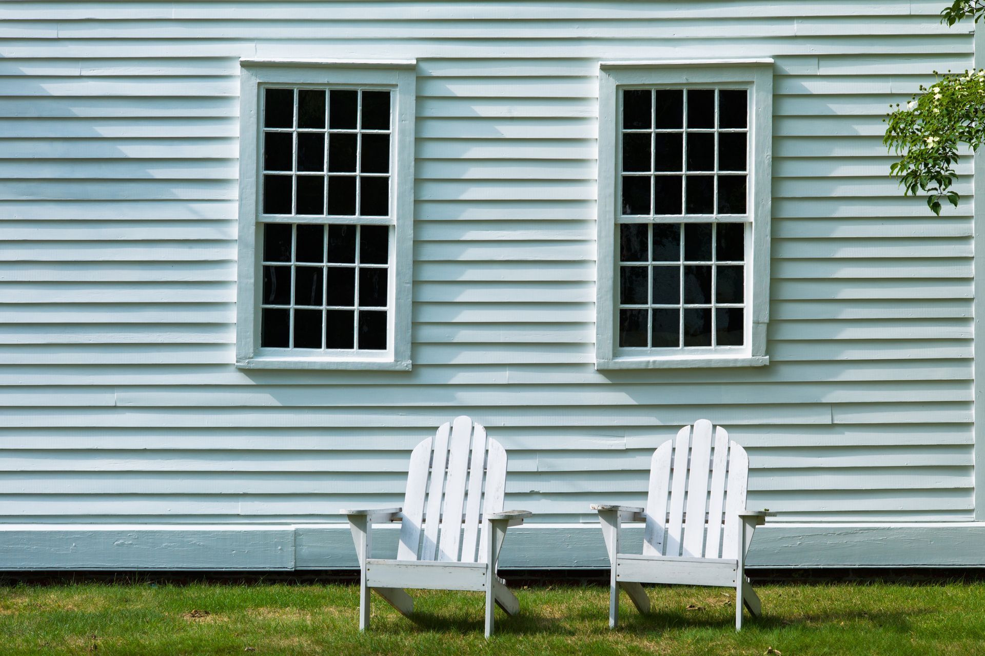 Two white Adirondack chairs on grass in front of a white house with two windows.