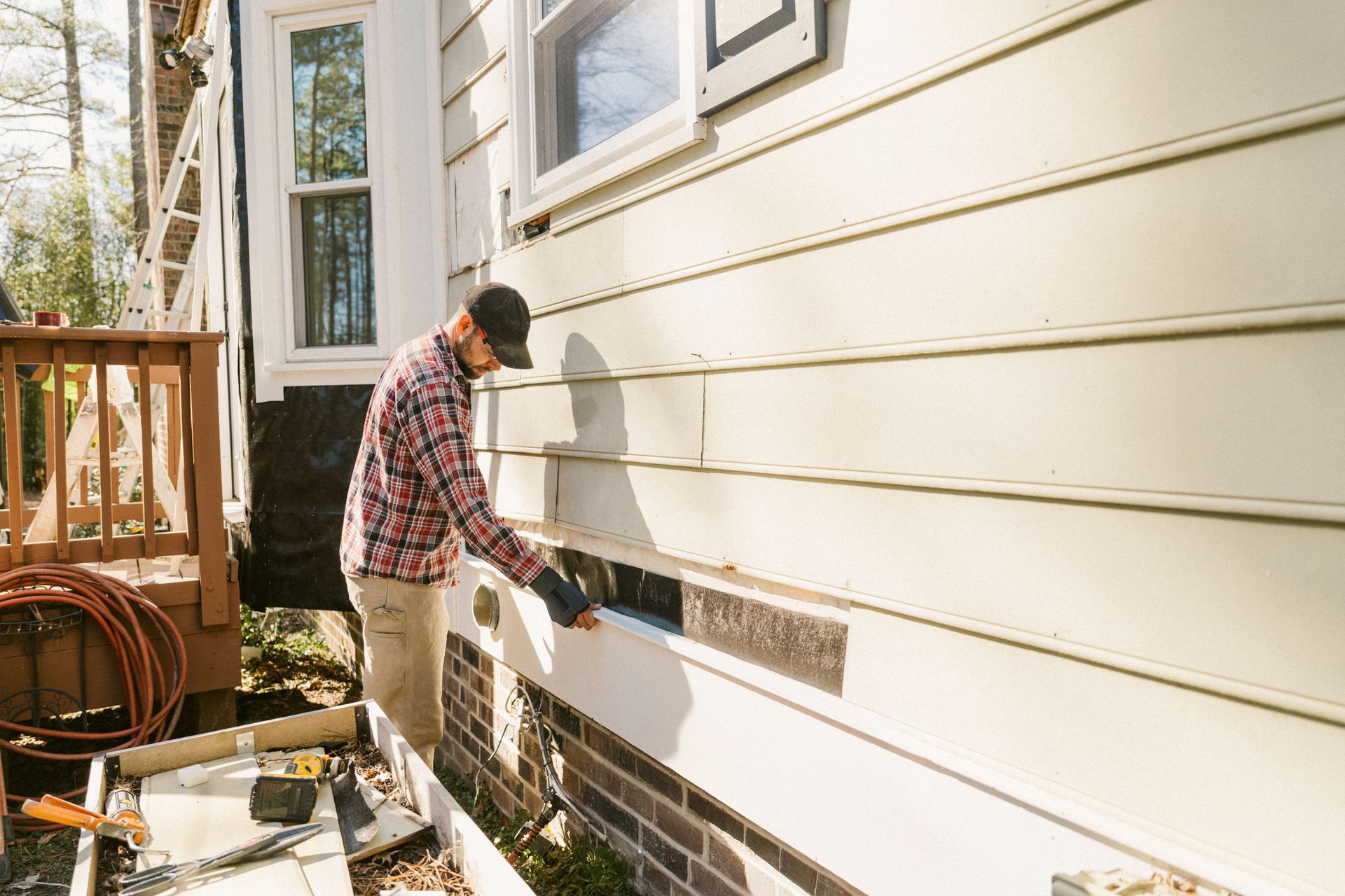 A man installing siding on a house exterior, near a window and deck, wearing a hat, gloves and plaid shirt.