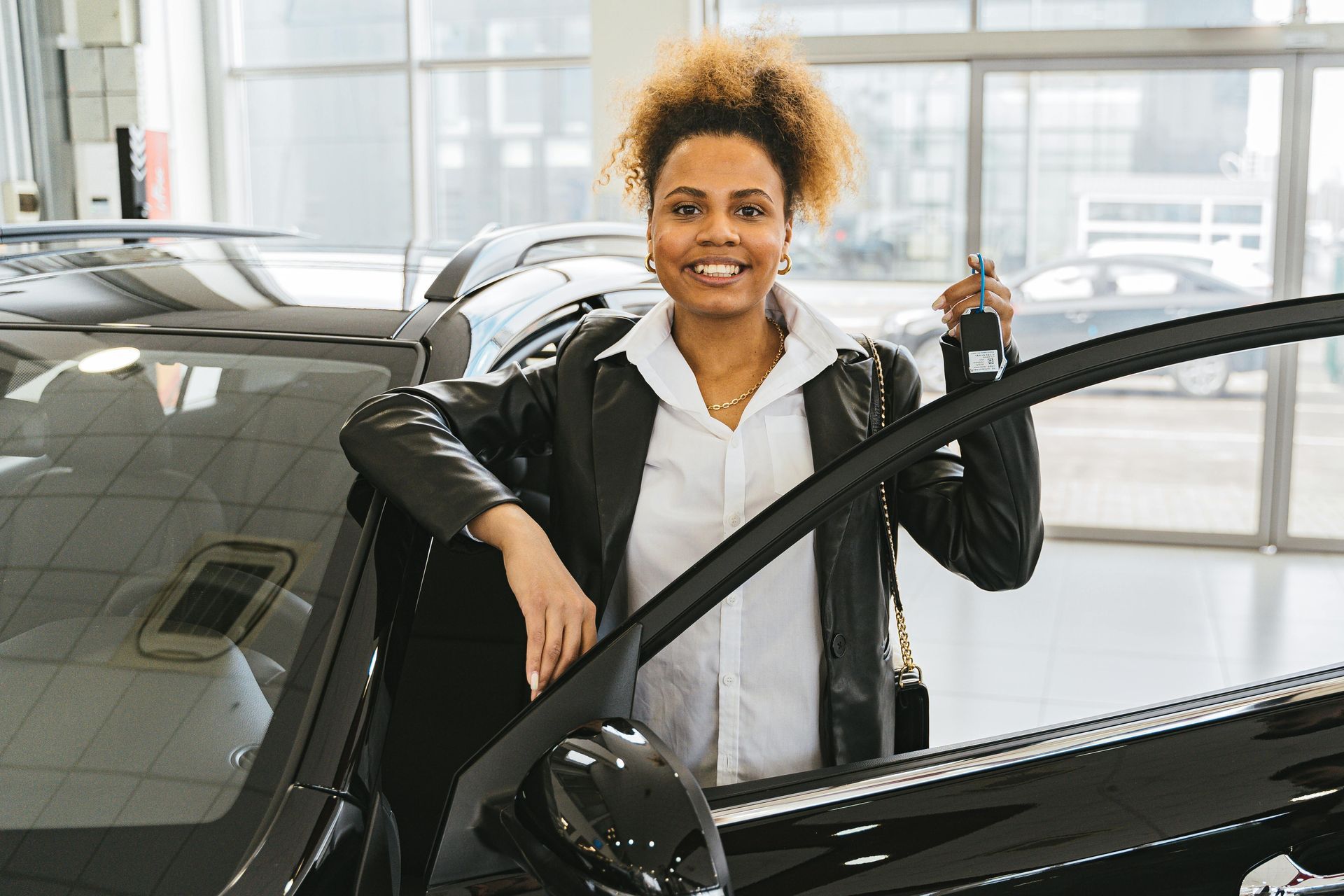 A woman is standing next to a car in a showroom holding a car key.
