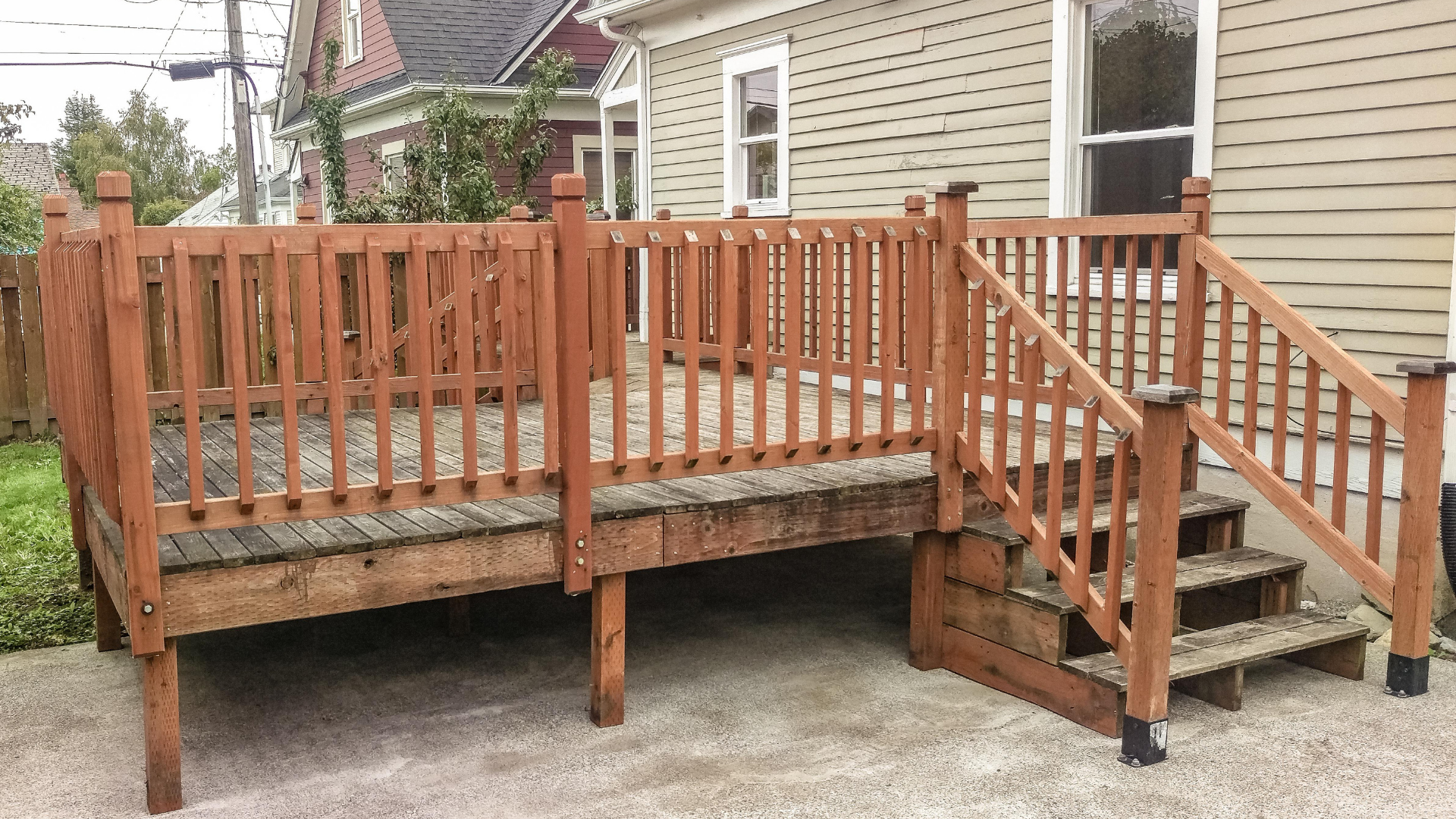 Wooden deck with stairs, railing, and posts next to a light-colored house.