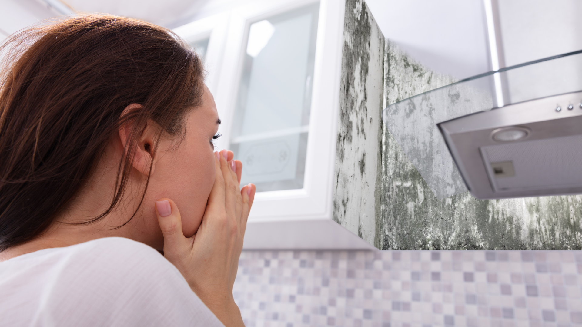 A woman is covering her face in a kitchen with mold on the cabinets.