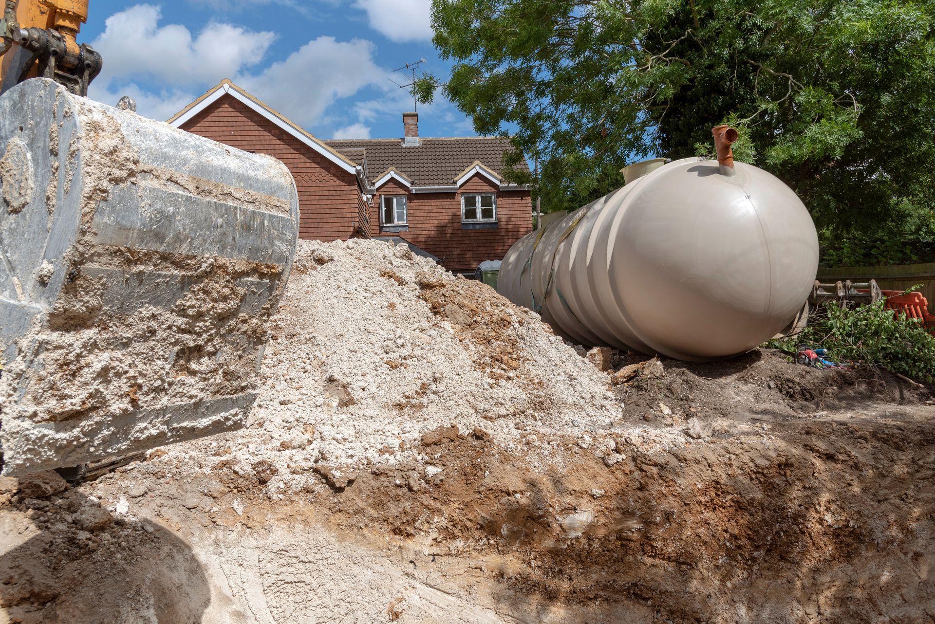 A large beige tank lies on a dirt construction site near a pile of soil and an excavator bucket, with a house in the back. A large beige tank lies on a dirt construction site near a pile of soil and an excavator bucket, with a house in the back.