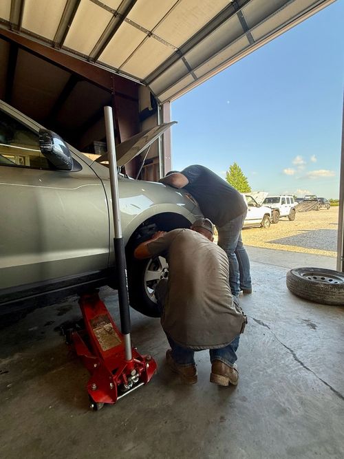 Two people work on the wheel of a silver vehicle elevated by a red hydraulic jack inside an open-air garage.