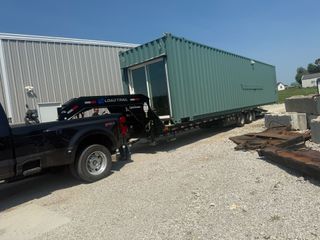 A black pickup truck towing a flatbed trailer loaded with a green shipping container on a gravel lot.