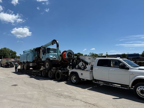 A light blue vacuum truck loaded onto a flatbed trailer being towed by a white pickup truck in a sunny, paved lot.