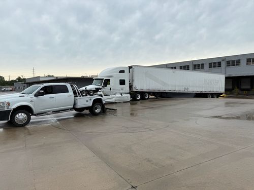 A white tow truck is towing a large white semi-truck and trailer in a paved industrial parking lot under a cloudy sky.