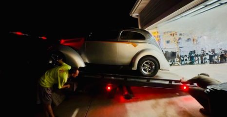 Two people assist in loading a vintage silver car onto a trailer parked outside a brightly lit garage at night.