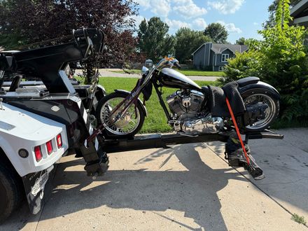 A custom motorcycle in black and silver is secured to the back of a white tow truck on a residential street.