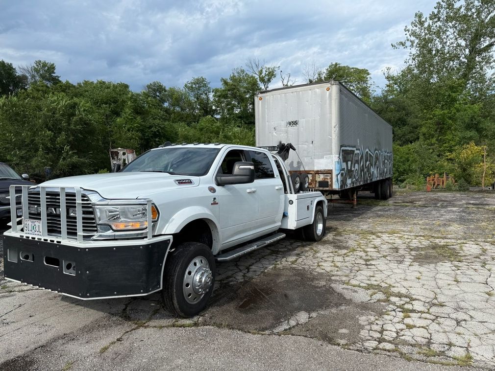 A white Ram dually pickup truck with a large black grill guard is hitched to a semi-trailer in an outdoor lot.