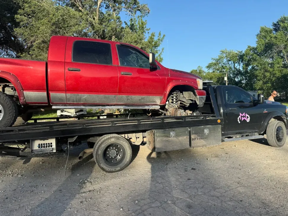 A red pickup truck is loaded onto a flatbed tow truck parked on a gravel surface under a clear blue sky.