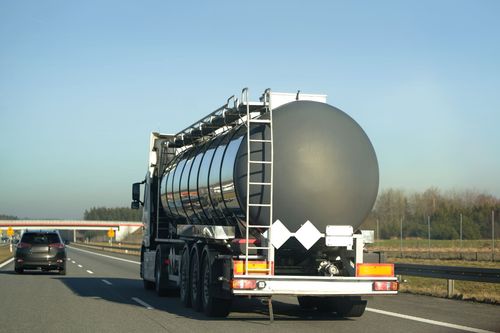 A silver tanker truck drives along a sunny highway, with another car traveling in the lane to its left.