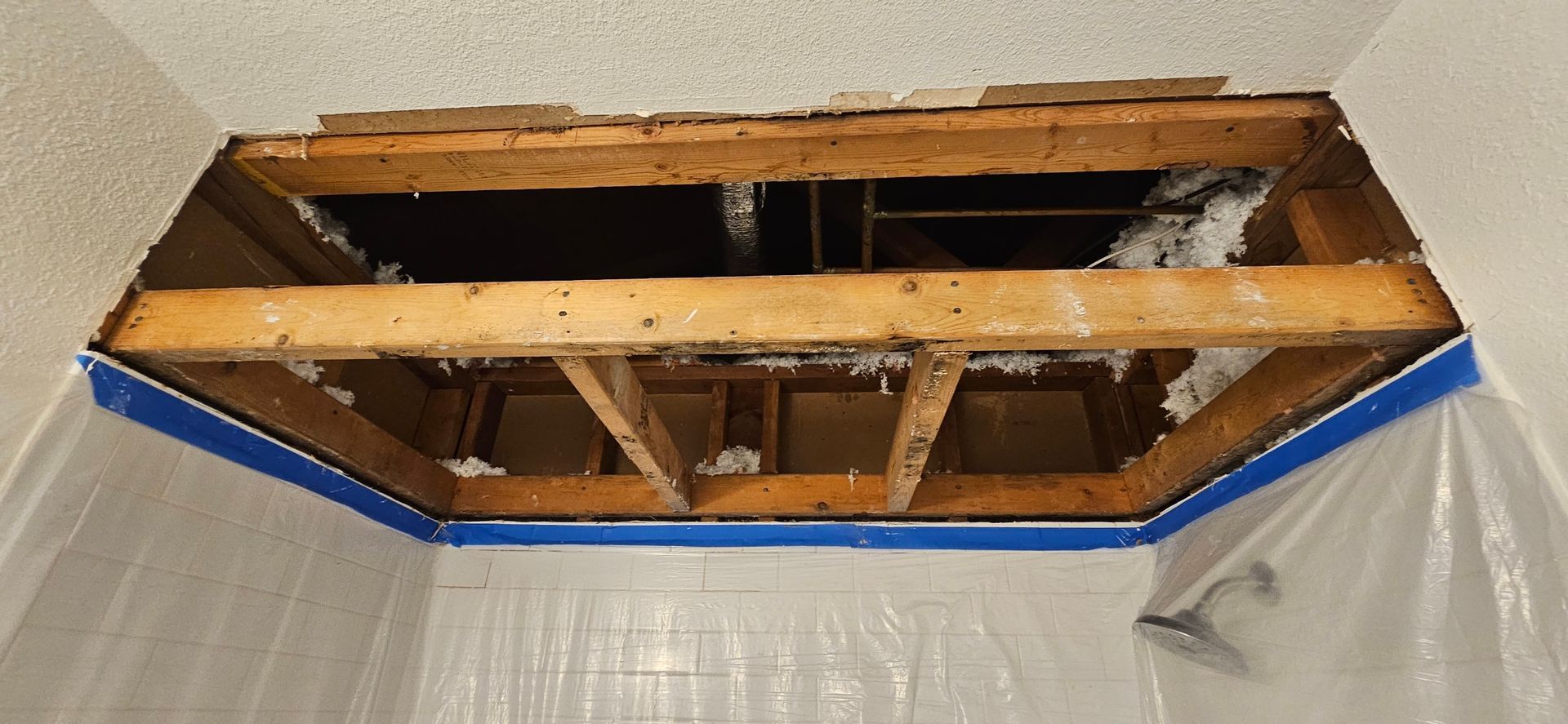 A hole in the bathroom ceiling reveals wooden beams, insulation, and the shower head. Blue tape lines the edges.