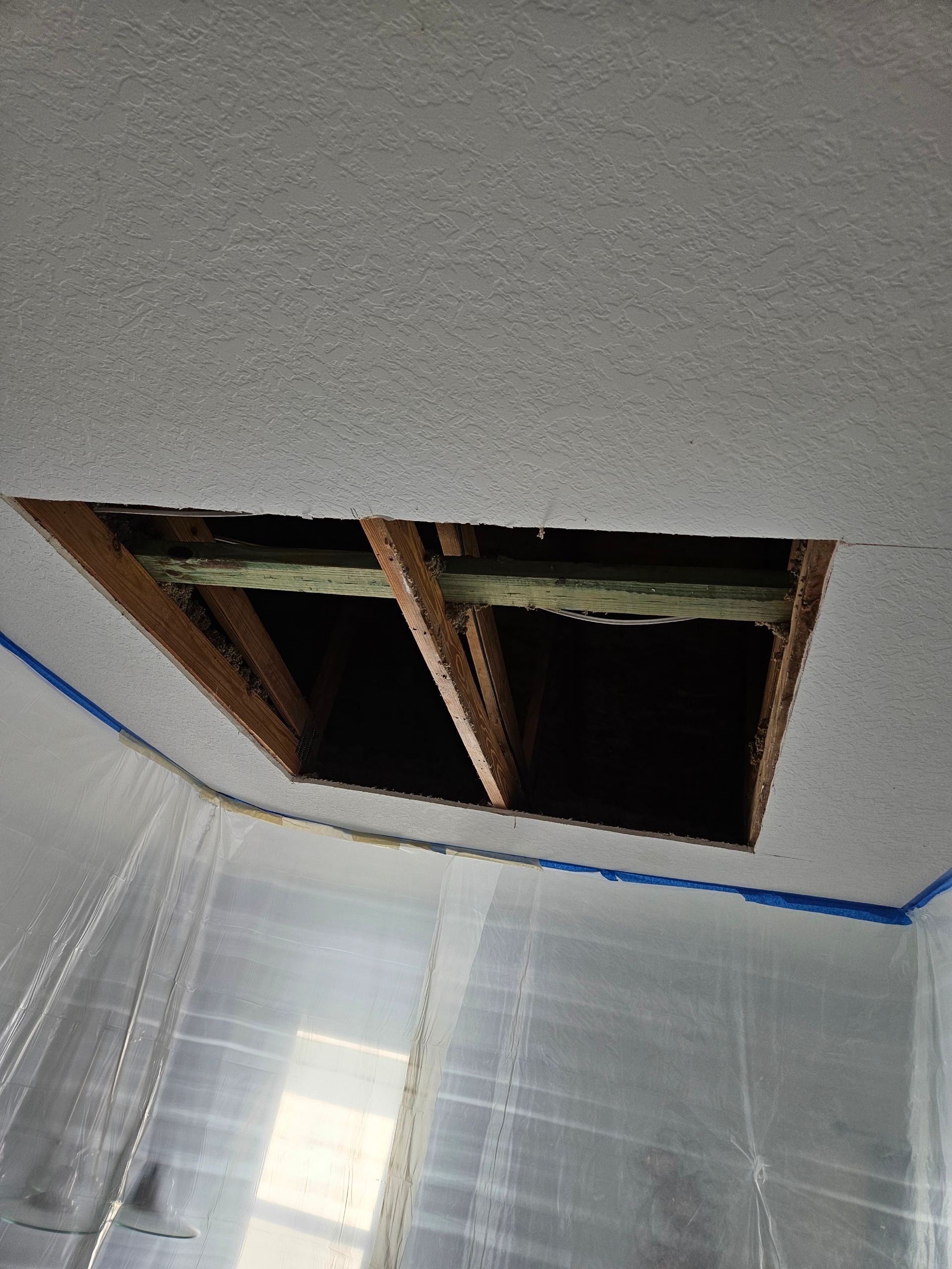 Hole in a textured white ceiling with exposed wooden beams, looking up from below.