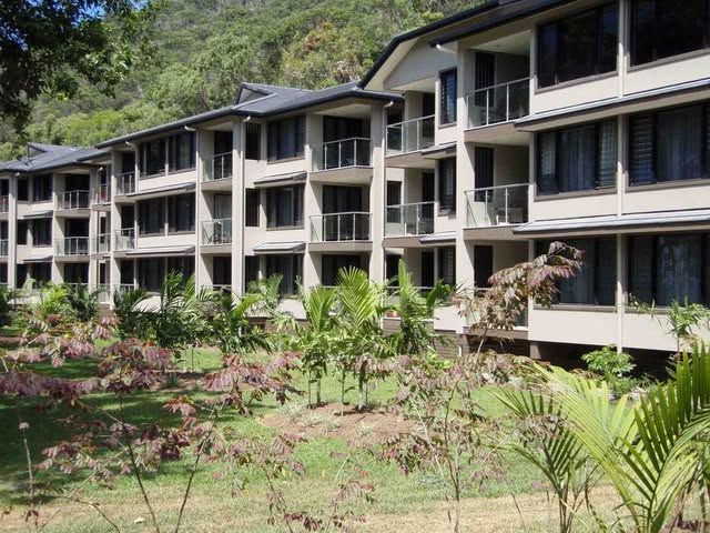 Modern apartment buildings with balconies beside a lush garden and hillside greenery