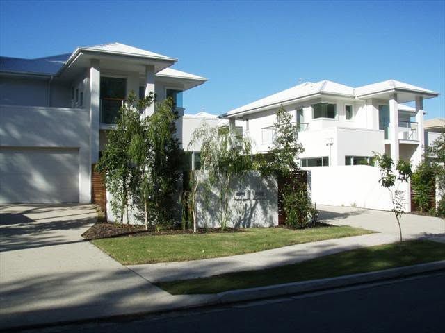 Modern white apartment buildings with balconies and a small landscaped lawn beside a road.