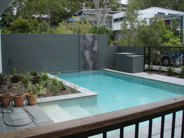 Modern backyard pool with turquoise water, potted plants, gray walls, and a wooden railing in the foreground