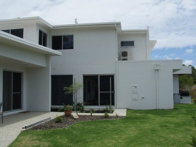 Modern white two-story house with a lawn, patio doors, and small landscaped garden under a cloudy sky