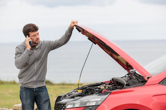 A man is talking on a cell phone while looking under the hood of a broken down car.