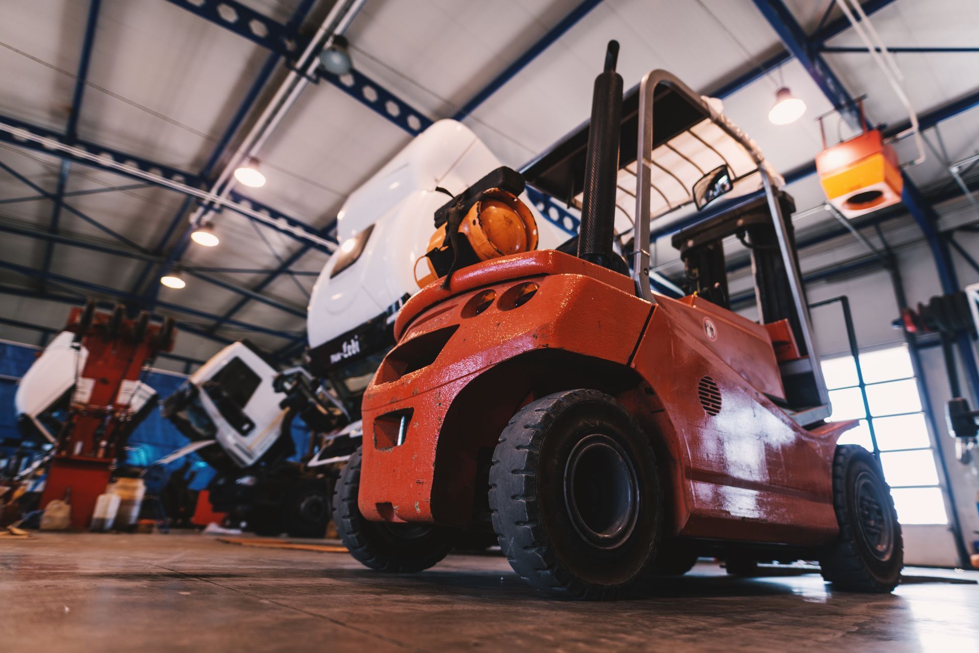 An orange forklift parked in a bright, industrial warehouse space with white trucks visible in the background.