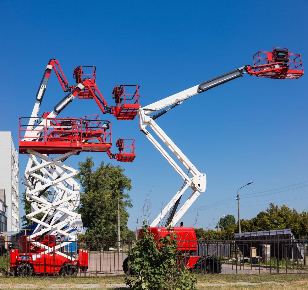 Two red and white aerial work platforms, one scissor lift and one boom lift, parked outdoors against a clear blue sky.