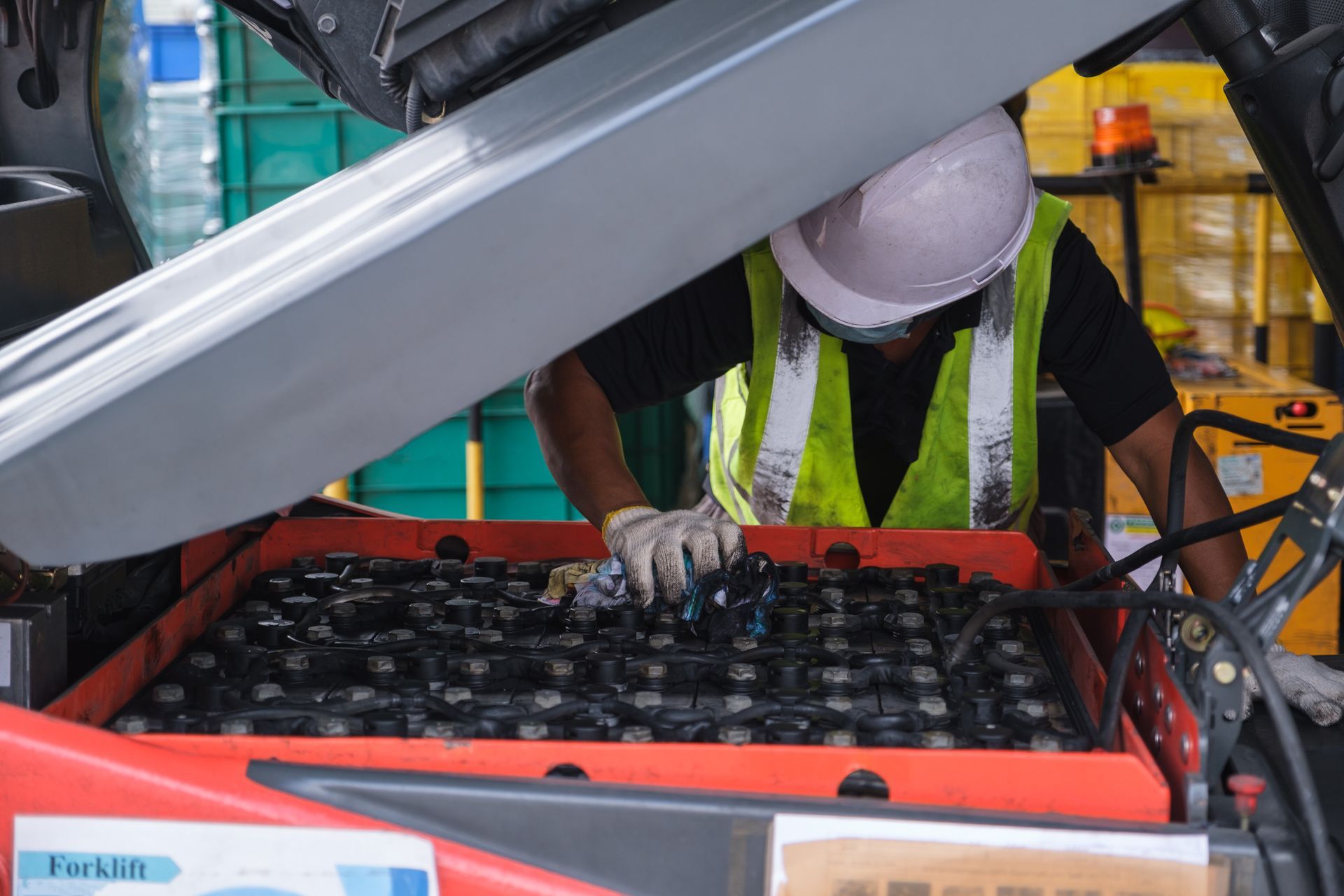 A worker in a high-visibility vest and hard hat performs maintenance on an open battery compartment of a forklift.