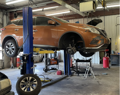 An orange SUV raised on a blue hydraulic lift inside an auto repair shop with one front wheel removed | Pro-Tec Auto Care