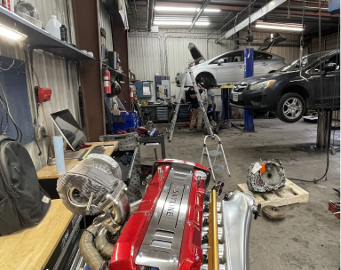 An engine with a red valve cover sits in the foreground of a mechanic's workshop with two cars lifted on auto lifts | Pro-Tec Auto Care